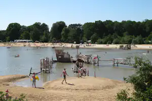 Blick auf den See mit Sandstrand und Stegen, Menschen am Ufer und im Wasser, Bäume am Rand.