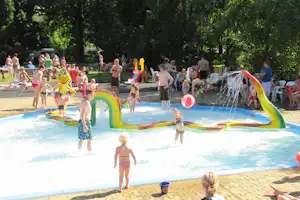 Kinder und Familien spielen im Campingpool mit bunten Wasserspielzeugen, im Hintergrund Bäume und Liegen am Beckenrand.