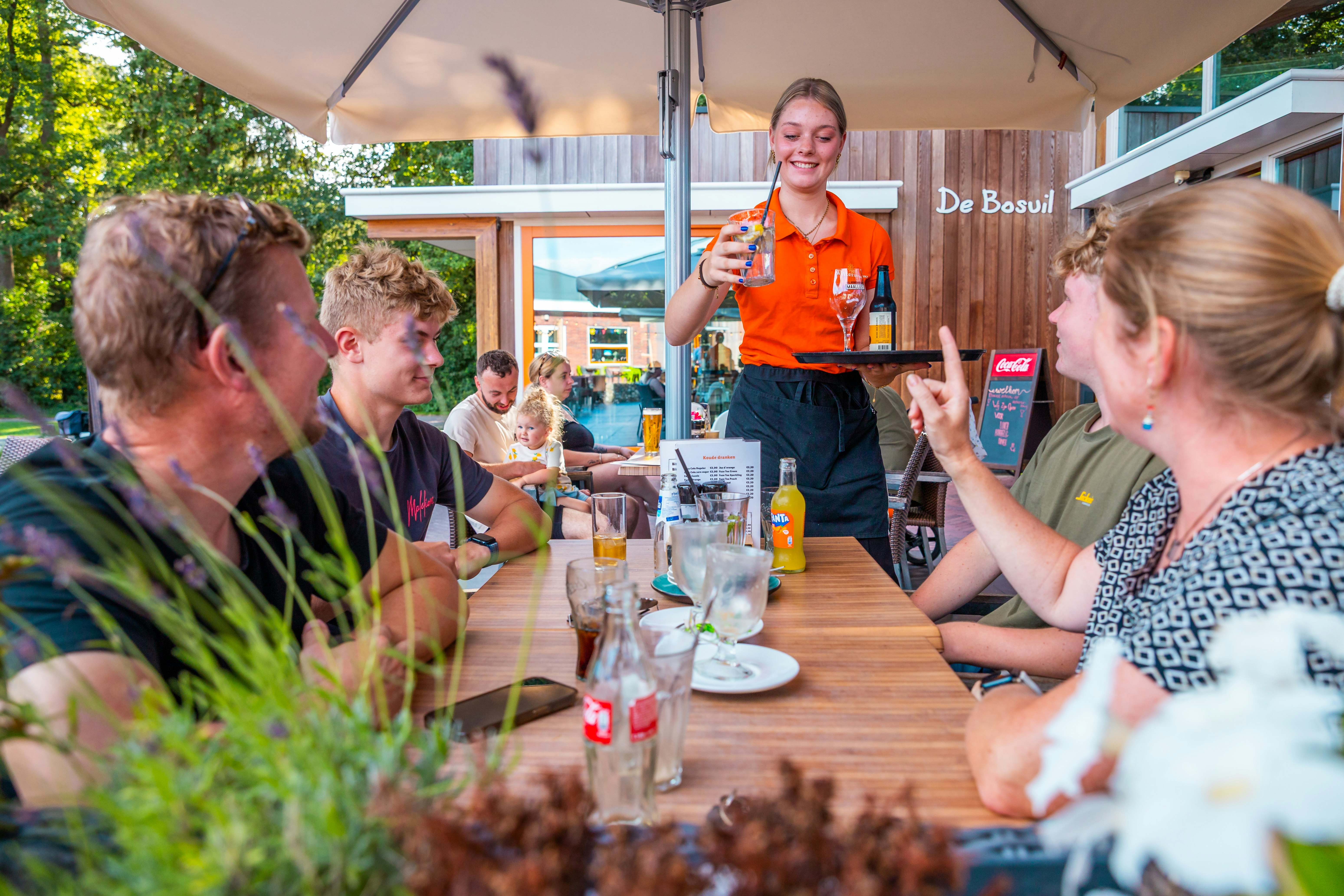 Gruppe beim Essen an einem gedeckten Tisch unter einem Zelt, Bedienung im orangefarbenen Shirt serviert Getränke im Außenbereich.