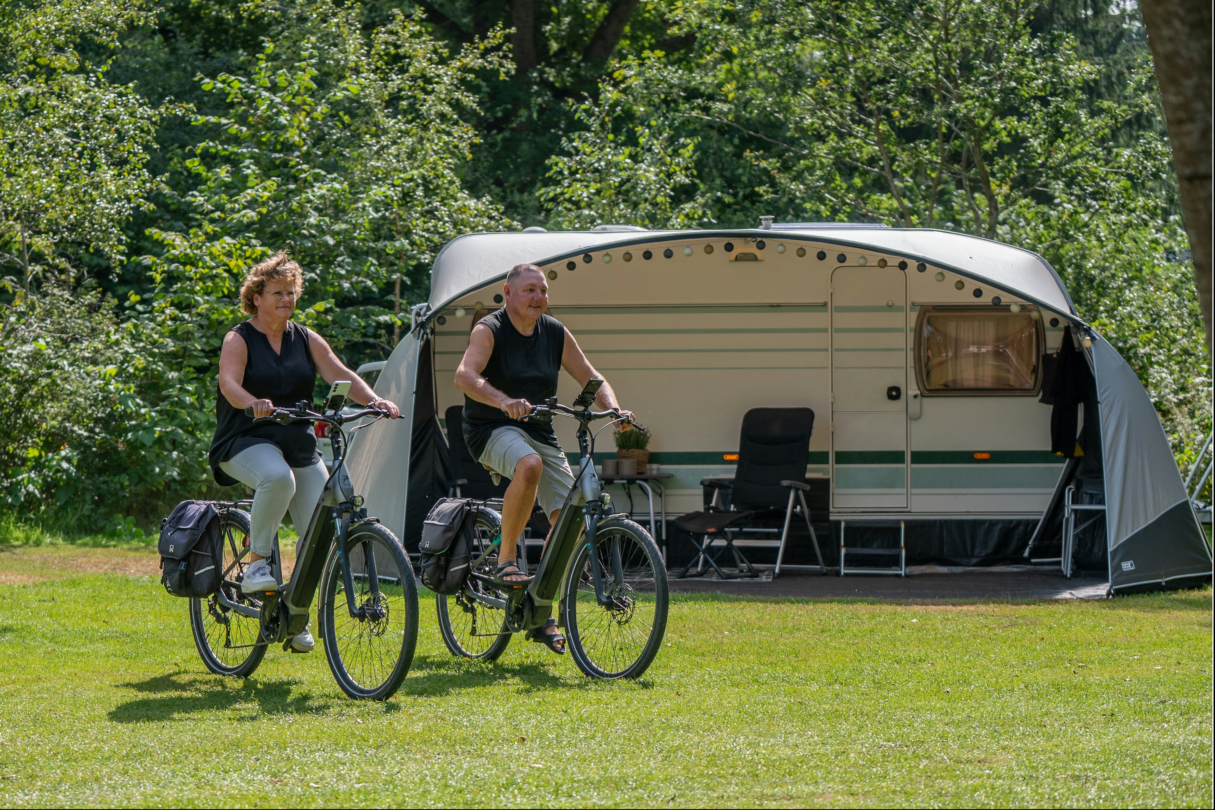 Zwei Personen mit Fahrrädern vor einem Wohnwagen-Zelt auf einem Campingplatz, umgeben von Rasen und Bäumen.
