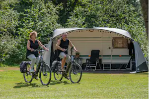 Zwei Personen mit Fahrrädern vor einem Wohnwagen-Zelt auf einem Campingplatz, umgeben von Rasen und Bäumen.