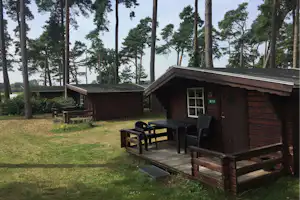 Holzhütte mit Terrasse und Sitzgruppe am Campingplatz im Wald, Rasenfläche und Bäume im Hintergrund.