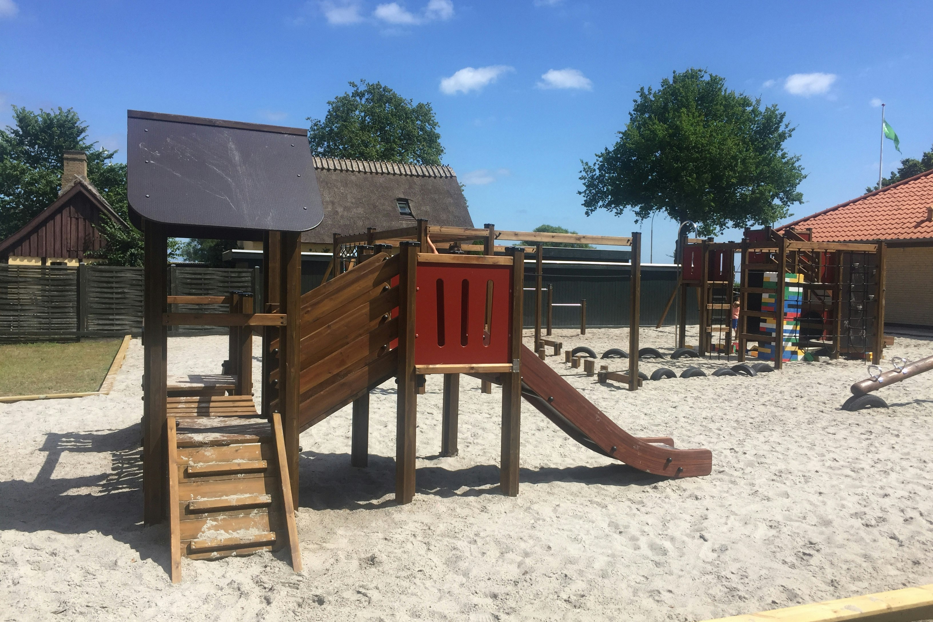 Spielplatz am Campingplatz mit Holzrutsche, Sandboden und Spielgeräten, im Hintergrund Gebäude und Bäume.