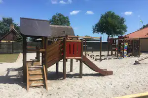 Spielplatz am Campingplatz mit Holzrutsche, Sandboden und Spielgeräten, im Hintergrund Gebäude und Bäume.