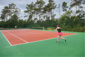 Tennisplatz auf einem Campingplatz mit rotem Boden, grüner Umrandung und Bäumen im Hintergrund.