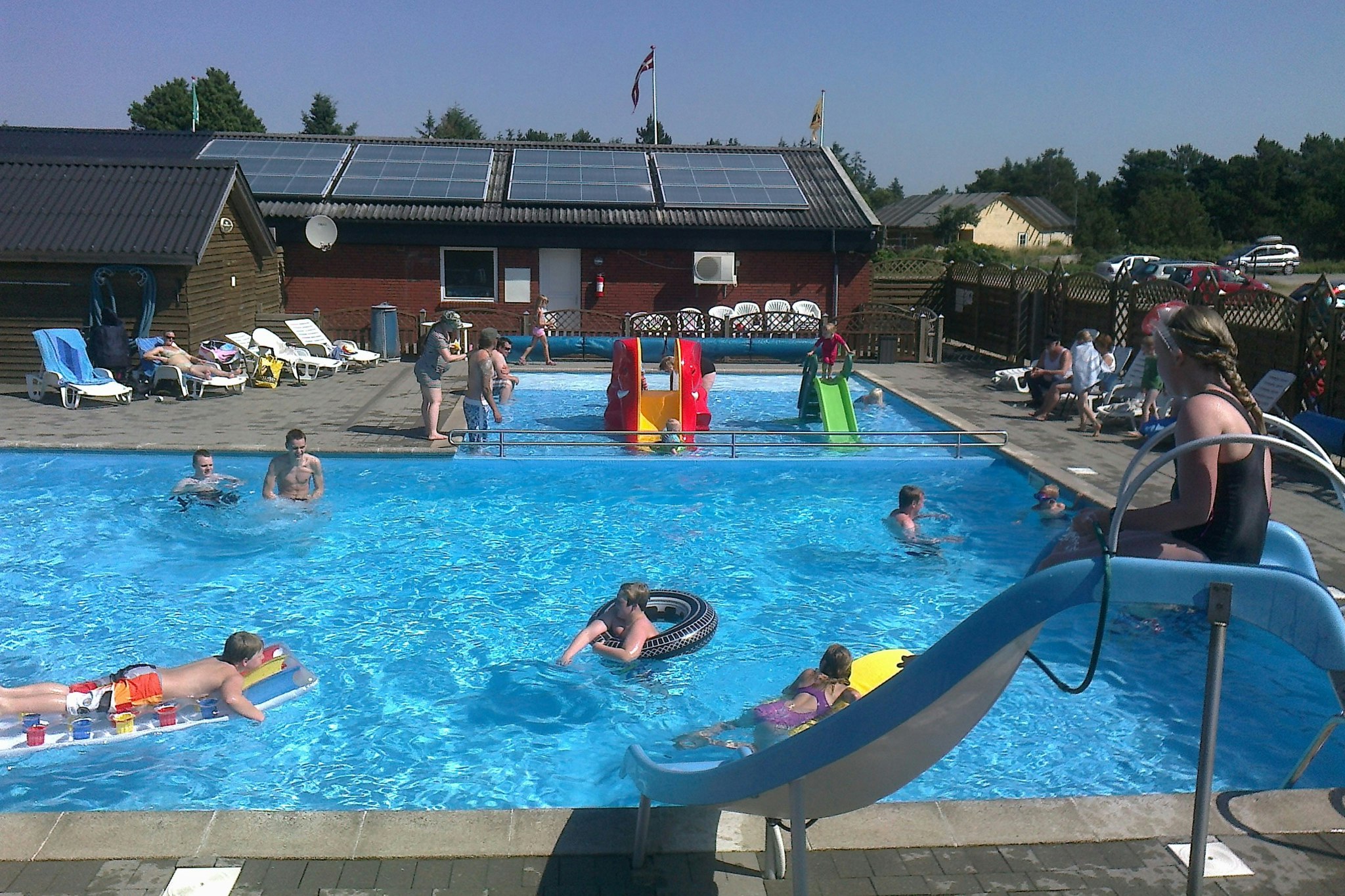 Campingplatz-Pool mit mehreren Liegestühlen, Menschen im Wasser und einem Gebäude mit Sonnenschirmen im Hintergrund.