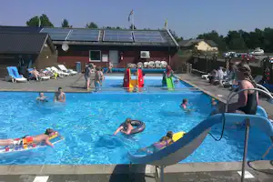 Campingplatz-Pool mit mehreren Liegestühlen, Menschen im Wasser und einem Gebäude mit Sonnenschirmen im Hintergrund.