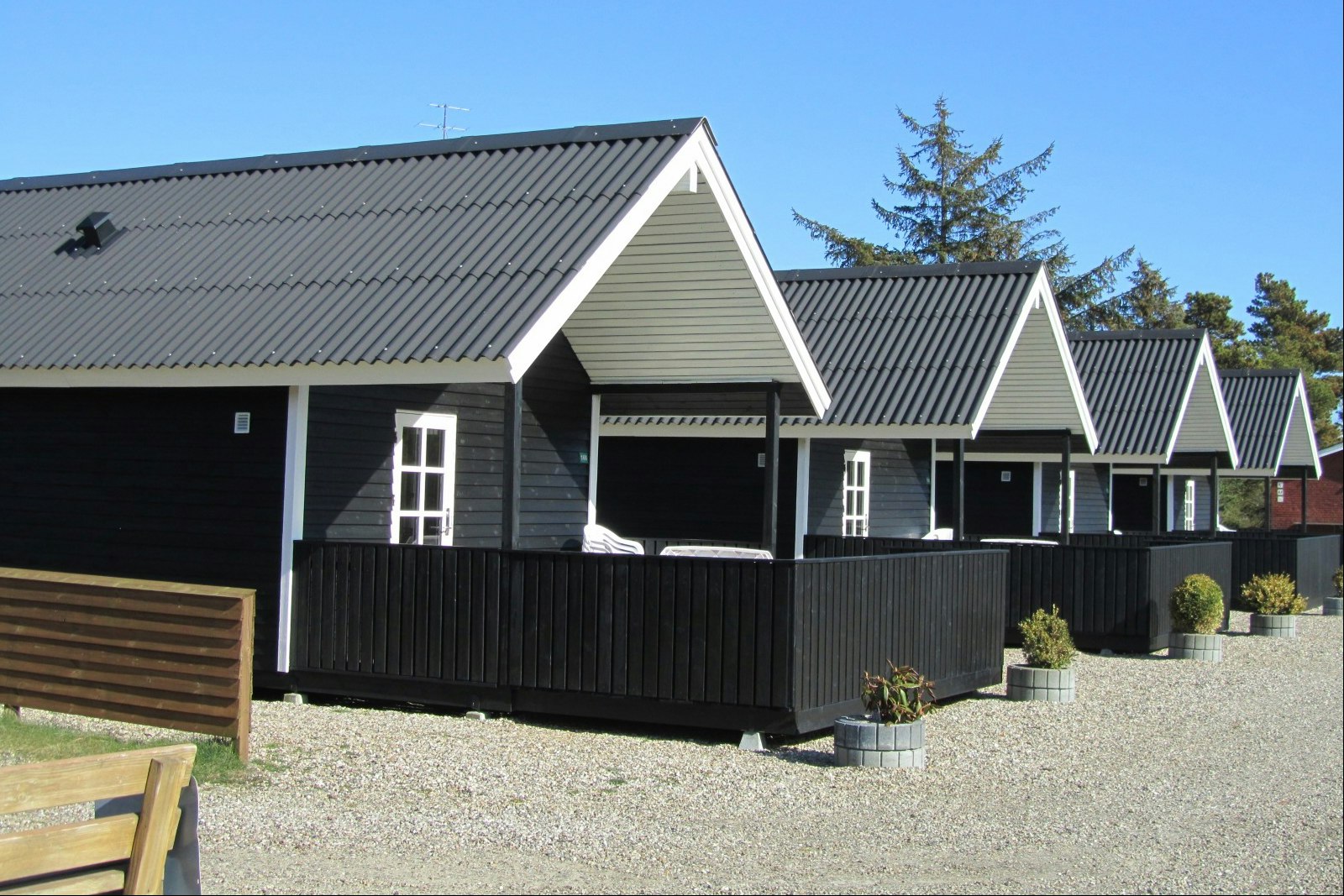 Reihen von Ferienhaeusern mit Holzfassade und schwarzen Dachern, davor ein Zaun und Kiesweg am Campingplatz.