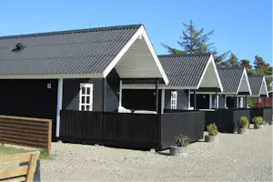 Reihen von Ferienhaeusern mit Holzfassade und schwarzen Dachern, davor ein Zaun und Kiesweg am Campingplatz.
