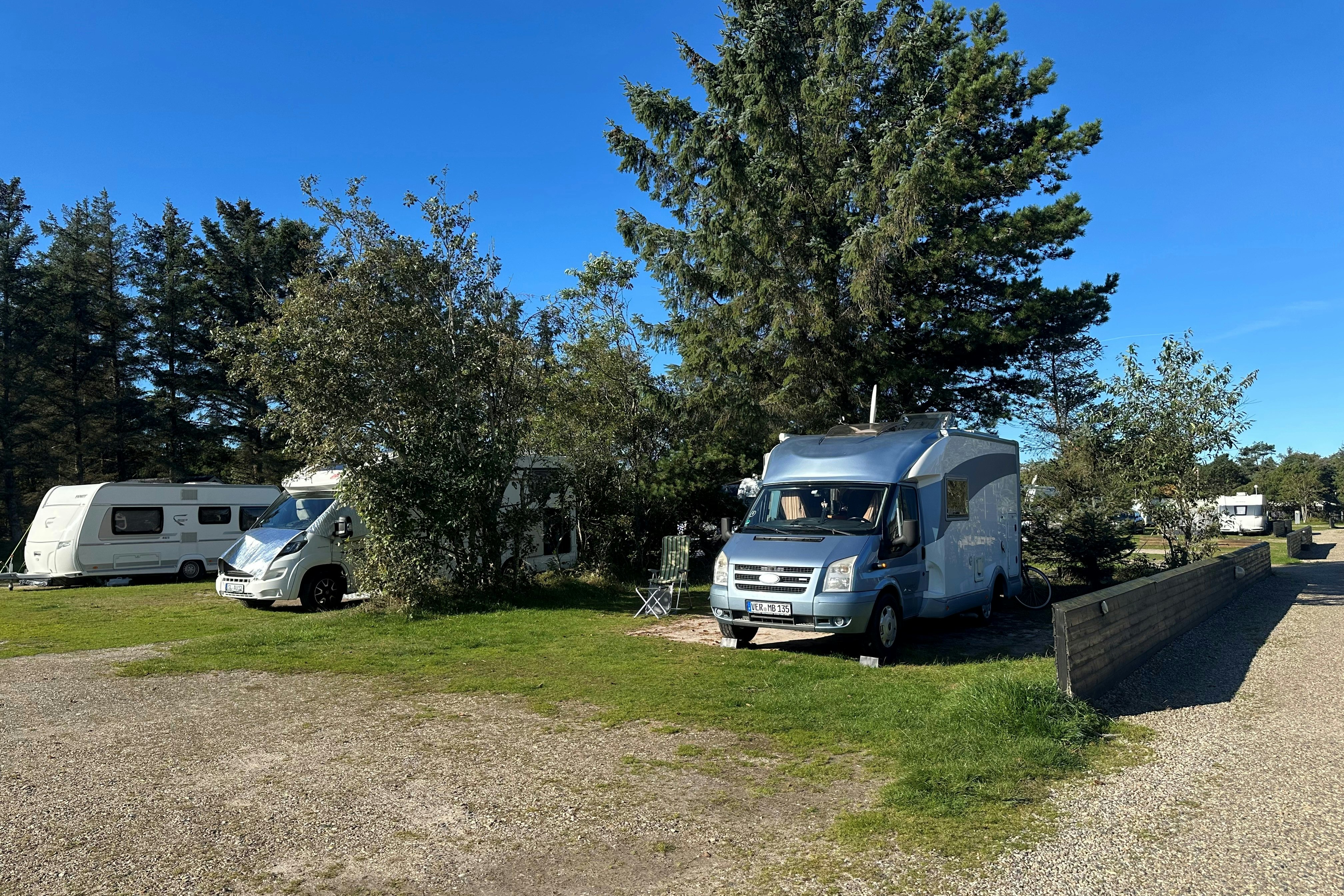 Wohnmobil und Auto auf einem Campingplatz, mit Kiesweg, Rasenfläche und Hecke, im Hintergrund weitere Fahrzeuge und Bäume.