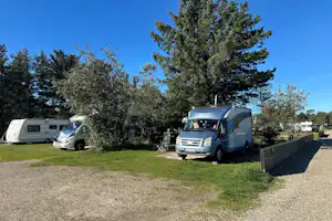 Wohnmobil und Auto auf einem Campingplatz, mit Kiesweg, Rasenfläche und Hecke, im Hintergrund weitere Fahrzeuge und Bäume.