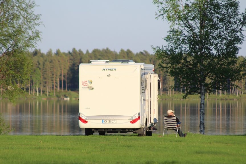 Wohnwagen am Seeufer im Grünen, dahinter Bäume und ein kleiner Tisch mit Stühlen.