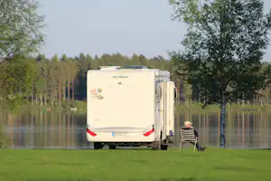 Wohnwagen am Seeufer im Grünen, dahinter Bäume und ein kleiner Tisch mit Stühlen.