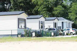 Weiße Mobilheime mit Fenstern und Schiebeläden, davor geparkte Autos und ein grüner Zaun, umgeben von Bäumen.