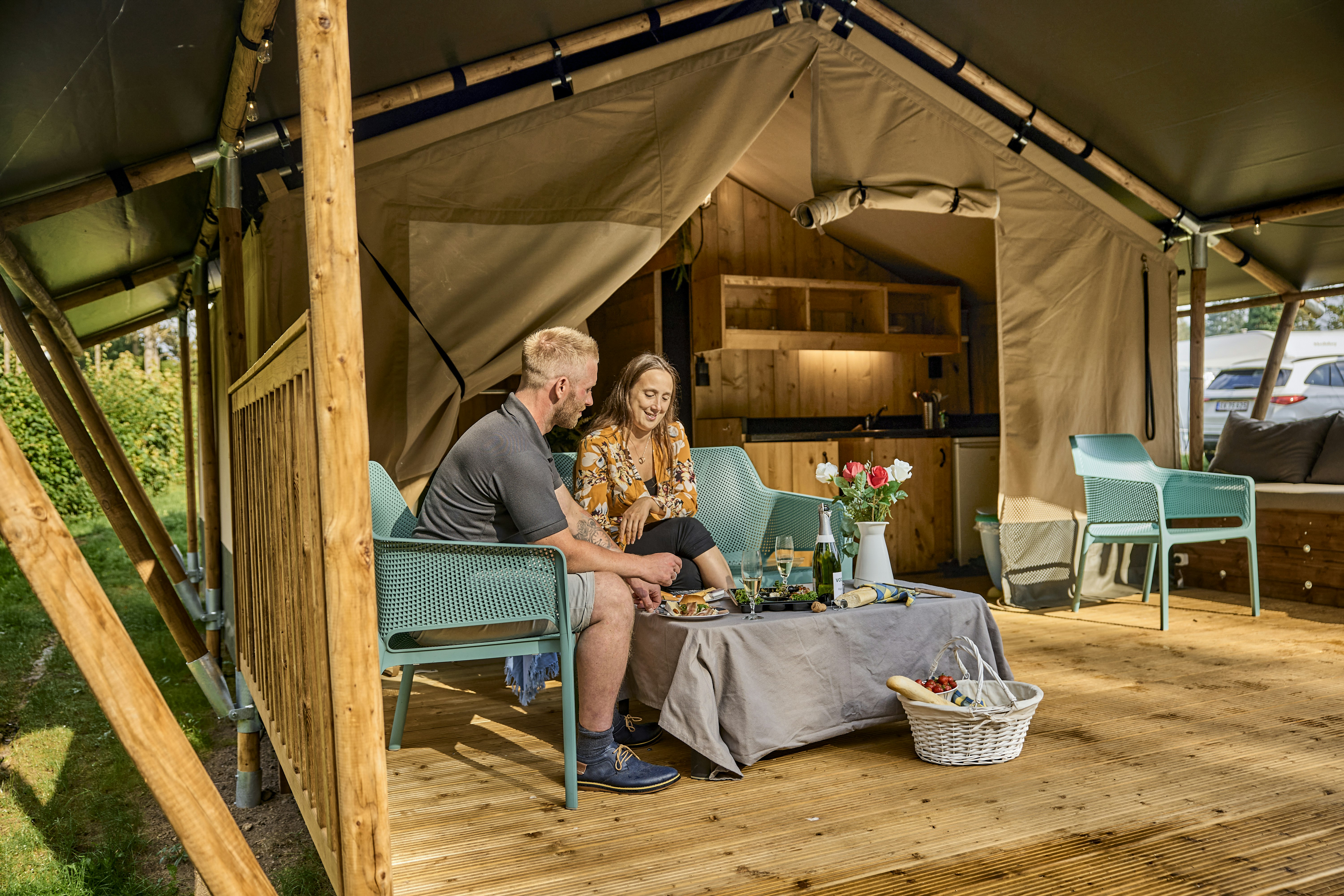 Zwei Personen sitzen am Campingtisch unter einem Zelt mit Holzrahmen, daneben Stühle, Blumen und eine Schüssel auf dem Boden.