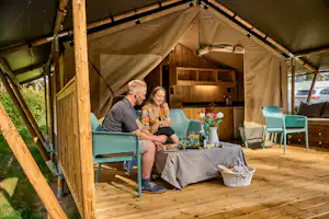 Zwei Personen sitzen am Campingtisch unter einem Zelt mit Holzrahmen, daneben Stühle, Blumen und eine Schüssel auf dem Boden.