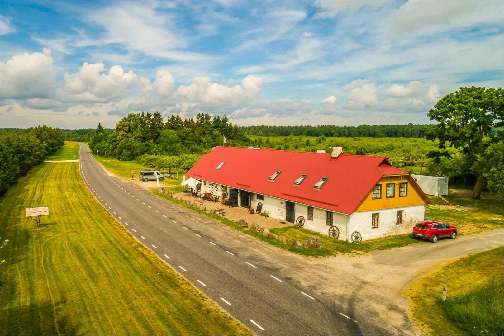 Landstraße am Campingplatz mit Rasenflächen, rotem Dachgebäude und geparktem Auto, umgeben von Bäumen und grünem Hügel.
