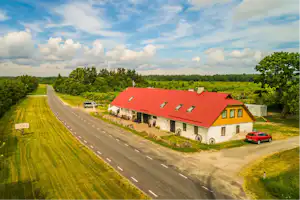 Landstraße am Campingplatz mit Rasenflächen, rotem Dachgebäude und geparktem Auto, umgeben von Bäumen und grünem Hügel.