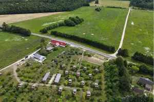Luftaufnahme eines Campingplatzes mit kleinen Stellplätzen, Wegen und Bäumen, umgeben von grünen Wiesen und einem Fluss im Hintergrund.
