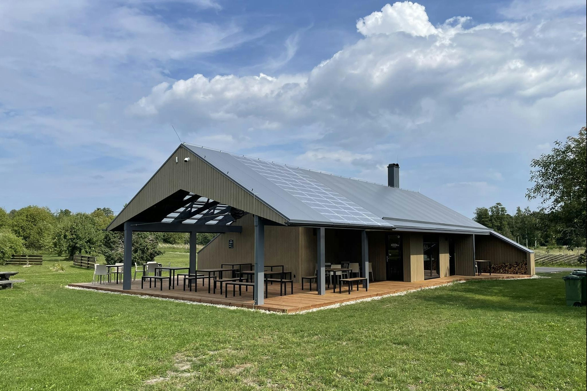 Großes Campinghaus mit überdachter Terrasse, Holzboden und Sitzgelegenheiten, umgeben von Rasen und Bäumen.