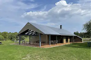 Großes Campinghaus mit überdachter Terrasse, Holzboden und Sitzgelegenheiten, umgeben von Rasen und Bäumen.