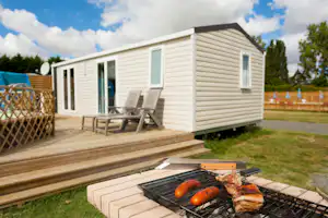 Holzterrasse mit Grill und Grillgut vor einem weißen Mobilheim, daneben Stühle und Rasenfläche mit Hecke und Bäumen.