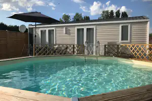 Swimmingpool mit klarem Wasser vor einer Terrasse am Mobilheim, mit Geländer, Holzsteg und weißen Wohnwänden.