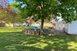 Campinggarten mit Rasen, Baum, Holzterrasse und Sitzbank; im Hintergrund ein Wohnwagen und ein Zaun.
