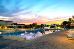 Schwimmbad mit Beckenrand, Liegen und Sonnenschirmen, im Hintergrund Gebäude und Bäume bei Sonnenuntergang.