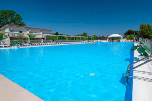 Blauer Campingpool mit Liegen, Sonnenschirm und Absperrgeländer; im Hintergrund Gebäude und Bäume.