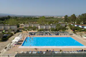 Blaue Schwimmbeckenanlage mit Liegestühlen, dahinter mehrstöckige Gebäude und Bäume in einer Campinganlage.