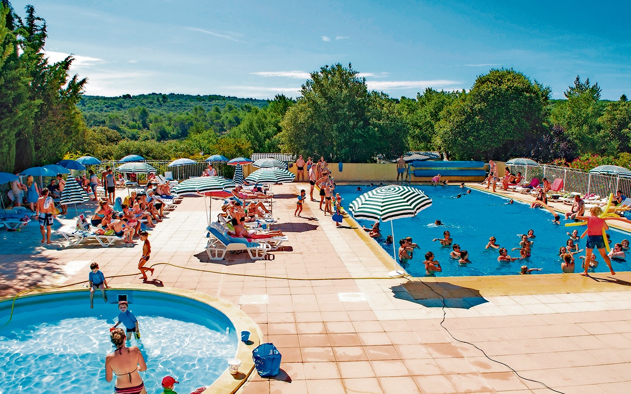 Großer Campingpool mit Kinderbereich, Liegen und Sonnenschirmen, daneben ein zweiter Pool und Wege zwischen Bäumen und Hügeln.
