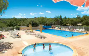 Camping-Pool mit blauem Wasser, vielen Liegen und Sonnenschirmen, umgeben von Bäumen und einem umzäunten Bereich.