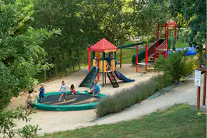 Spielplatz mit rotem Klettergerüst, Rutsche und Sandkasten, umgeben von Bäumen und Hecken am Campingplatz.