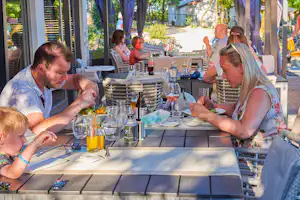Familie am gedeckten Tisch unter Sonnenschirmen im Campingrestaurant, Gläser und Getränke auf dem Tisch, Kinder und Erwachsene.