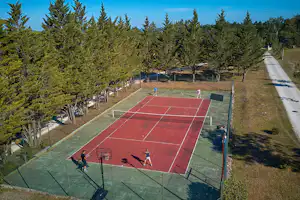 Roter Tennisplatz im Campinggelände, umgeben von hohen Nadelbäumen, mit Zaun, Wegen und mehreren Spielbereichen.