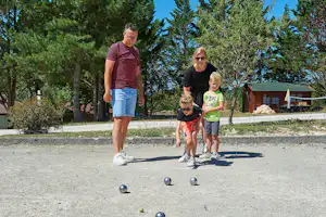 Mann und Kinder spielen Boule auf einem Campingplatzweg, im Hintergrund Bäume und ein Wohnwagen.