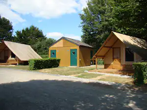 Holzhütten und Zelte auf einem Campingplatz, mit grünem Eingangstor, Hecken und Wegen zwischen den Unterkünften.