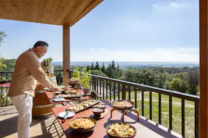 Mann grillt auf dem Camping-Balkon an einem gedeckten Tisch, mit Holzüberdachung und weitem Blick ins Tal.