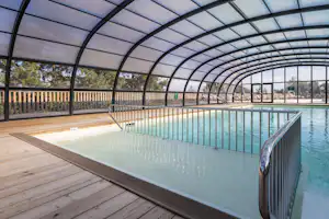 Überdachter Pool mit Holzterrasse, Geländer und Liegen im Hintergrund unter einem Glasdach.