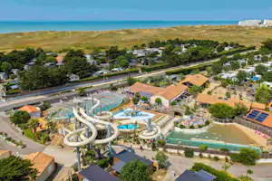 Luftaufnahme eines Campingplatzes mit mehreren Pools, Wasserrutschen und Wegen, umgeben von Bäumen und Häusern nahe dem Meer.