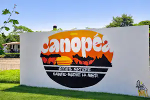 Camping-Eingangsschild mit Aufdruck „Janopere“ und „Gîtes Nature“ vor Rasen, Bäumen und Gebäuden.
