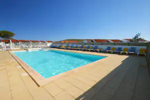 Schwimmbad mit türkisfarbenem Wasser, Steinboden und Liegen am Rand, dahinter Reihen von Ferienhäusern mit roten Dächern.