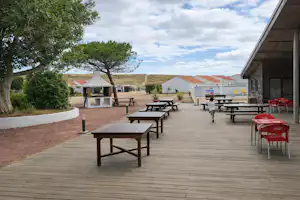 Holzterrasse mit mehreren Tischen und Stühlen, daneben ein Gebäude, im Hintergrund Zelte und Bäume.