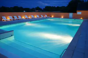 Beleuchteter Campingpool am Abend mit Liegen auf der Terrasse und dunklen Bäumen im Hintergrund.