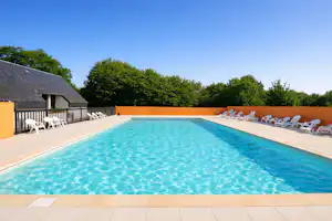 Schwimmbecken auf dem Campingplatz mit Liegestühlen am Rand, gelbem Gebäude und Bäumen im Hintergrund.