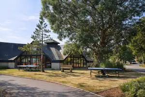 Campinghaus mit Terrasse, Holzbänken und Tisch, daneben ein Weg; im Hintergrund Bäume und ein weiteres Gebäude.
