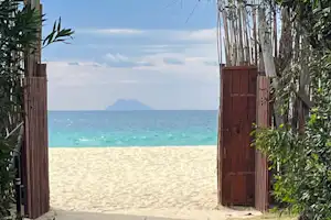 Blick durch einen Gartenweg mit Holztoren auf den Sandstrand und das türkisfarbene Meer, im Hintergrund ein Berg.