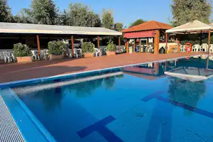 Schwimmbecken im Campingplatz mit Sonnenschirmen, Liegen und Tischen, im Hintergrund ein Gebäude mit Ziegeldach und Bäumen.