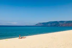 Weicher Sandstrand mit Blick auf das türkisfarbene Meer und eine kleine Gruppe am Ufer, im Hintergrund Hügel und Himmel.